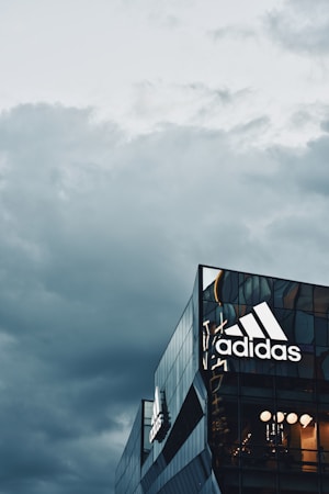 A modern building with reflective glass panels features a prominent Adidas logo. The architecture is sleek and contemporary, with parts of the building's interior visible through the windows. Overhead, a cloudy sky casts a moody atmosphere.