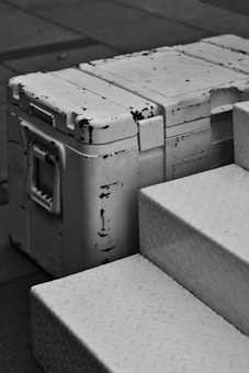 A vintage metal box with a weathered, distressed look sits next to metal steps with a patterned surface. The box has noticeable black marks and scratches, and the surroundings imply an industrial or urban setting.