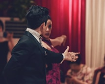 A couple engaged in an intimate and passionate dance, with the man in a formal black suit and the woman in a vibrant red dress. The background features red curtains, and other people are slightly out of focus, possibly watching the performance.