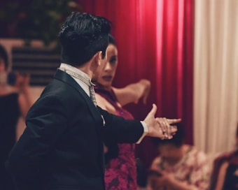 A couple engaged in an intimate and passionate dance, with the man in a formal black suit and the woman in a vibrant red dress. The background features red curtains, and other people are slightly out of focus, possibly watching the performance.