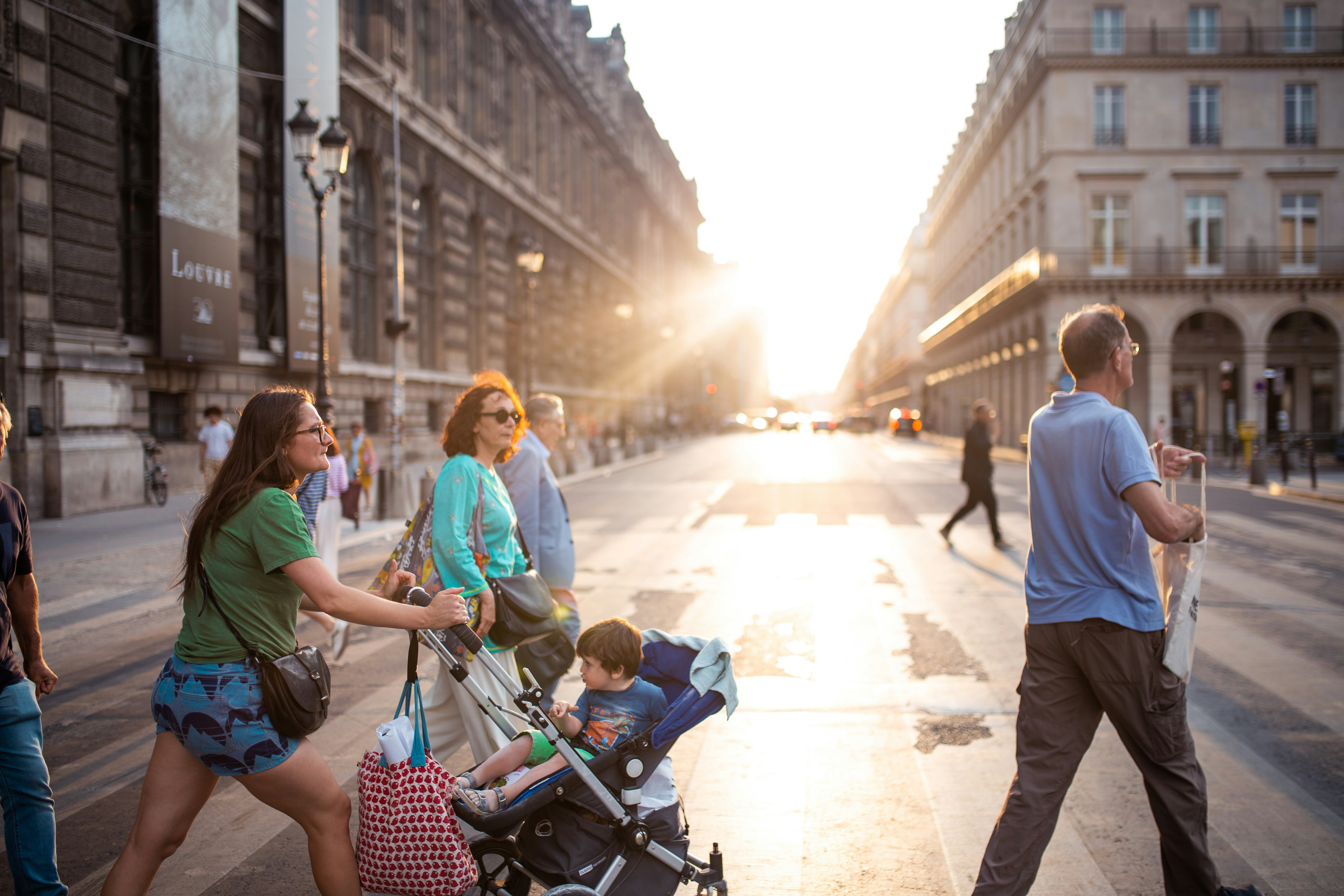Mother pushing a toddler in a lightweight stroller across a sunlit Paris street, with shoppers crossing beside her