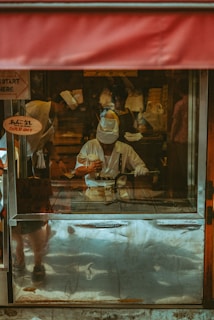 A person dressed in a white uniform and hat is engaging in culinary preparation inside a food stall. The setup includes a metal cooking surface with visible utensils. A red awning is partially visible above, and signs in a foreign language appear on the stall's glass panels.