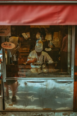 A person dressed in a white uniform and hat is engaging in culinary preparation inside a food stall. The setup includes a metal cooking surface with visible utensils. A red awning is partially visible above, and signs in a foreign language appear on the stall's glass panels.