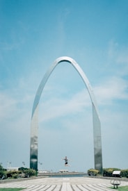 A person is captured mid-jump in front of a large, metallic, arched structure set against a clear blue sky. The scene is expansive, with the arch dominating the middle of the image and the person positioned centrally, highlighted by the bright daylight. Surrounding the arch, there are patches of greenery and a few buildings in the distance.