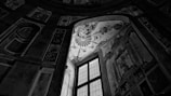 A monochrome interior scene featuring a large, intricately decorated wall with a high window. The artwork on the walls includes ornate patterns and baroque style frescoes, illuminated by light filtering through the window.