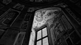 A monochrome interior scene featuring a large, intricately decorated wall with a high window. The artwork on the walls includes ornate patterns and baroque style frescoes, illuminated by light filtering through the window.