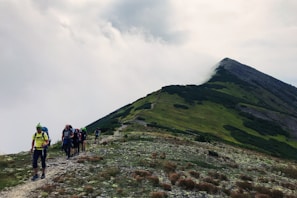 A group of hikers is walking up a rugged mountain path covered in rocks and sparse vegetation. The mountain is partially shrouded in clouds, creating a dramatic backdrop. The sky is overcast, adding a moody atmosphere to the scene.