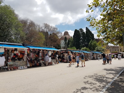 A bustling outdoor market with multiple stalls selling various colorful goods. People are walking around, exploring the merchandise under a partly cloudy sky. The market is set against a backdrop of trees and a historical dome structure visible in the distance.