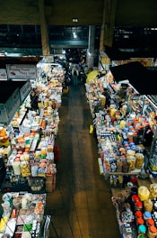 An overhead view of a bustling indoor market with stalls on either side filled with various goods. The stalls are packed with colorful packages, jars, and containers, suggesting a variety of snacks, spices, and dried foods. People are seen walking through the narrow aisles between the stalls, indicating a vibrant and busy atmosphere.