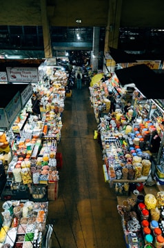 An overhead view of a bustling indoor market with stalls on either side filled with various goods. The stalls are packed with colorful packages, jars, and containers, suggesting a variety of snacks, spices, and dried foods. People are seen walking through the narrow aisles between the stalls, indicating a vibrant and busy atmosphere.