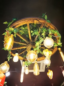 A rustic chandelier made from a wooden wagon wheel, adorned with numerous vintage-style filament light bulbs and entwined with green leafy vines. The setup hangs from a dark ceiling, creating a warm, inviting atmosphere.