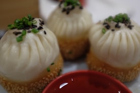 Three steamed buns topped with chopped green onions and black sesame seeds, resting on a bed of golden sesame seeds with a red bowl partially visible below.