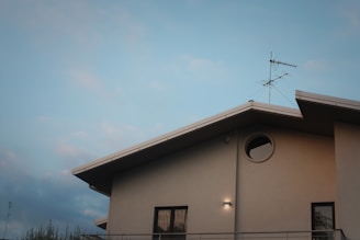 A modern building with clean architectural lines features a flat-roofed section and a circular window. An antenna is mounted on the roof against a clear blue sky.