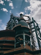 An industrial structure with large metal frameworks, pipes, and a towering chimney under a partly cloudy sky. The structure features intricate ladders and platforms with rusted and weathered surfaces.