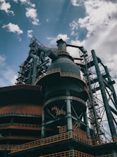 An industrial structure with large metal frameworks, pipes, and a towering chimney under a partly cloudy sky. The structure features intricate ladders and platforms with rusted and weathered surfaces.