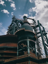 An industrial structure with large metal frameworks, pipes, and a towering chimney under a partly cloudy sky. The structure features intricate ladders and platforms with rusted and weathered surfaces.
