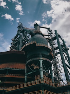 An industrial structure with large metal frameworks, pipes, and a towering chimney under a partly cloudy sky. The structure features intricate ladders and platforms with rusted and weathered surfaces.