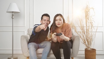 A man and a woman sitting on a light-colored sofa engaged in video gaming. The man, wearing a dark t-shirt and jeans, is pointing outward, while the woman, dressed in a brown shirt and dark pants, is holding a game controller. A tall lamp stands to the left, and a pot with decorative branches is on the right, with soft lighting enhancing the cozy and casual setting.