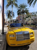 A vibrant yellow luxury convertible car is parked on a street lined with tall palm trees. The area appears upscale, with high-end brand stores visible in the background. The scene is well-lit by bright sunlight, casting sharp shadows on the sidewalk.