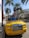 A vibrant yellow luxury convertible car is parked on a street lined with tall palm trees. The area appears upscale, with high-end brand stores visible in the background. The scene is well-lit by bright sunlight, casting sharp shadows on the sidewalk.