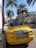 A vibrant yellow luxury convertible car is parked on a street lined with tall palm trees. The area appears upscale, with high-end brand stores visible in the background. The scene is well-lit by bright sunlight, casting sharp shadows on the sidewalk.