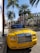 A vibrant yellow luxury convertible car is parked on a street lined with tall palm trees. The area appears upscale, with high-end brand stores visible in the background. The scene is well-lit by bright sunlight, casting sharp shadows on the sidewalk.