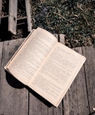 An open book rests on a wooden surface, with text in a language that uses Cyrillic script. The wooden background is weathered, and there is grass visible in the upper right corner, adding a touch of nature.