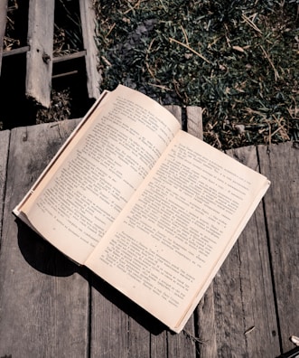 An open book rests on a wooden surface, with text in a language that uses Cyrillic script. The wooden background is weathered, and there is grass visible in the upper right corner, adding a touch of nature.