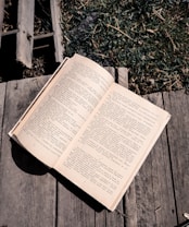 An open book rests on a wooden surface, with text in a language that uses Cyrillic script. The wooden background is weathered, and there is grass visible in the upper right corner, adding a touch of nature.