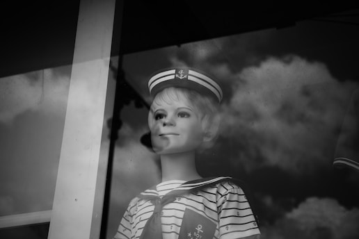 A vintage mannequin with a sailor outfit is seen behind a glass window. The mannequin wears a striped shirt and a sailor hat, and the reflection of clouds is visible on the glass, adding a dreamy effect. The black and white tone enhances the nostalgic and somewhat eerie feel of the scene.