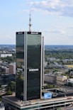 A tall, modern skyscraper with reflective glass panels is prominently displaying logos of large corporations. The building is surrounded by a sprawling urban landscape with lots of greenery, smaller buildings, and an expansive blue sky with scattered clouds.