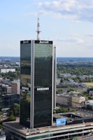 A tall, modern skyscraper with reflective glass panels is prominently displaying logos of large corporations. The building is surrounded by a sprawling urban landscape with lots of greenery, smaller buildings, and an expansive blue sky with scattered clouds.