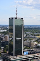 A tall, modern skyscraper with reflective glass panels is prominently displaying logos of large corporations. The building is surrounded by a sprawling urban landscape with lots of greenery, smaller buildings, and an expansive blue sky with scattered clouds.
