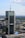 A tall, modern skyscraper with reflective glass panels is prominently displaying logos of large corporations. The building is surrounded by a sprawling urban landscape with lots of greenery, smaller buildings, and an expansive blue sky with scattered clouds.