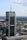 A tall, modern skyscraper with reflective glass panels is prominently displaying logos of large corporations. The building is surrounded by a sprawling urban landscape with lots of greenery, smaller buildings, and an expansive blue sky with scattered clouds.