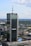 A tall, modern skyscraper with reflective glass panels is prominently displaying logos of large corporations. The building is surrounded by a sprawling urban landscape with lots of greenery, smaller buildings, and an expansive blue sky with scattered clouds.