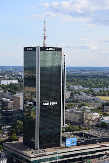 A tall, modern skyscraper with reflective glass panels is prominently displaying logos of large corporations. The building is surrounded by a sprawling urban landscape with lots of greenery, smaller buildings, and an expansive blue sky with scattered clouds.