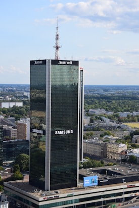 A tall, modern skyscraper with reflective glass panels is prominently displaying logos of large corporations. The building is surrounded by a sprawling urban landscape with lots of greenery, smaller buildings, and an expansive blue sky with scattered clouds.