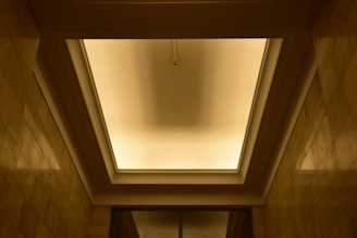 A recessed ceiling with a central light fixture casting a warm glow. The ceiling is framed by shadowed rectangular panels, and the walls are lined with stone tiles that reflect the soft lighting.