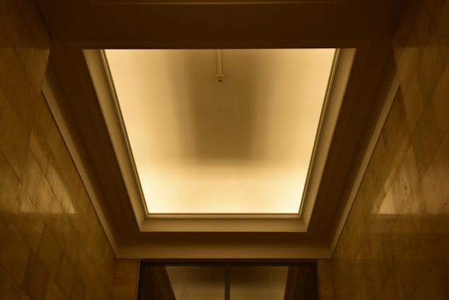 A recessed ceiling with a central light fixture casting a warm glow. The ceiling is framed by shadowed rectangular panels, and the walls are lined with stone tiles that reflect the soft lighting.
