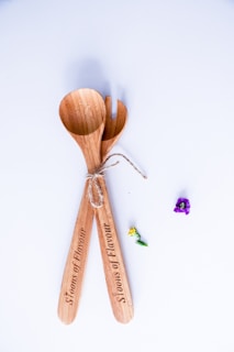 Two wooden salad utensils, tied together with a piece of twine, are placed on a light-colored surface. The utensils have the words 'Spoons of Flavour' engraved on their handles. Nearby, two small flowers, one purple and one yellow, add a touch of color.