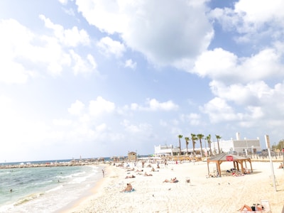 A sunny beach scene with numerous people relaxing on the sand and swimming in the sea. The beach is lined with beach huts and sun umbrellas, and several palm trees add a tropical atmosphere. The sky is partly cloudy with bright blue tones.