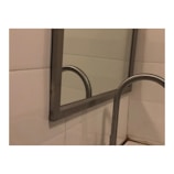 A metal framed mirror is mounted on a tiled wall, reflecting part of a metallic handrail. The handrail and part of the bathroom tiles are visible, with the scene suggesting a bathroom setting.