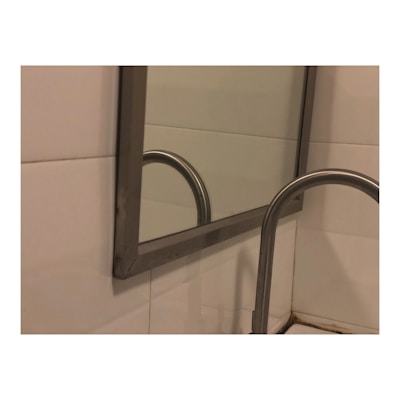 A metal framed mirror is mounted on a tiled wall, reflecting part of a metallic handrail. The handrail and part of the bathroom tiles are visible, with the scene suggesting a bathroom setting.