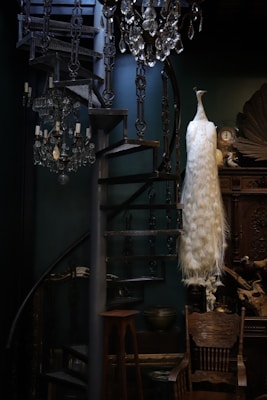 An elegantly decorated interior features a spiral staircase made of dark wood and metal with ornate designs. Two crystal chandeliers hang overhead, adding a touch of opulence. A striking white taxidermy peacock stands prominently on a wooden chair, its long plumage cascading down. The room includes intricate wooden furniture and various decorative items, creating a sense of vintage luxury.