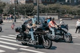 Two motorcyclists riding side by side on a city street, both wearing helmets and casual attire. One motorcycle has an American flag attached to the back. Pedestrians and other vehicles are visible in the background, and the scene is captured at an intersection with a zebra crossing.