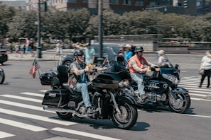 Two motorcyclists riding side by side on a city street, both wearing helmets and casual attire. One motorcycle has an American flag attached to the back. Pedestrians and other vehicles are visible in the background, and the scene is captured at an intersection with a zebra crossing.