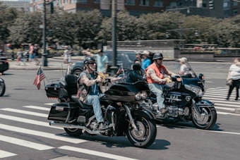Two motorcyclists riding side by side on a city street, both wearing helmets and casual attire. One motorcycle has an American flag attached to the back. Pedestrians and other vehicles are visible in the background, and the scene is captured at an intersection with a zebra crossing.