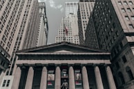 A neoclassical building with tall columns and an American flag atop its roof is framed by skyscrapers under a cloudy sky. The building has banners featuring historical figures and text on its facade.