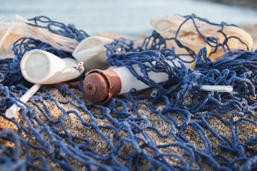 A sandy beach is littered with various types of plastic waste and blue fishing nets. Items such as plastic bottles, caps, and straws are entangled in the netting, highlighting pollution and environmental concerns.
